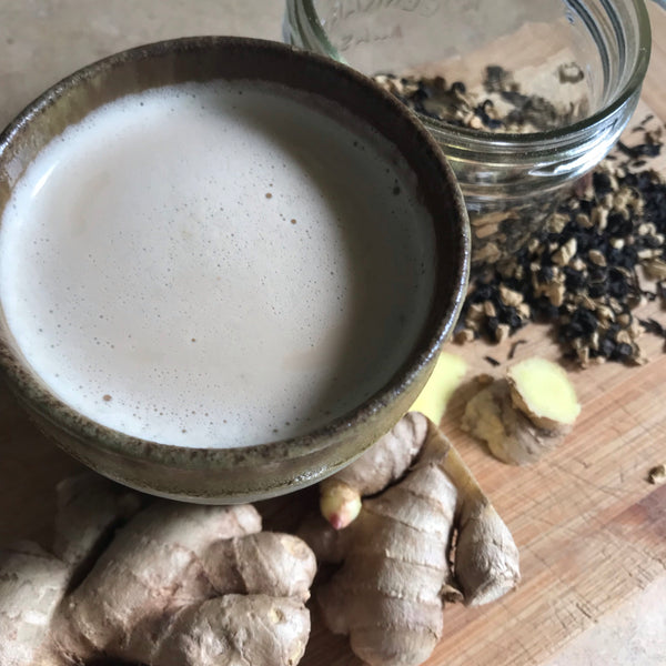 A cup of Shanti Chai & Co's Candied Ginger Chain on a wooden cutting board, surrounded by fresh ginger slices and a Mason jar full of chai spices.